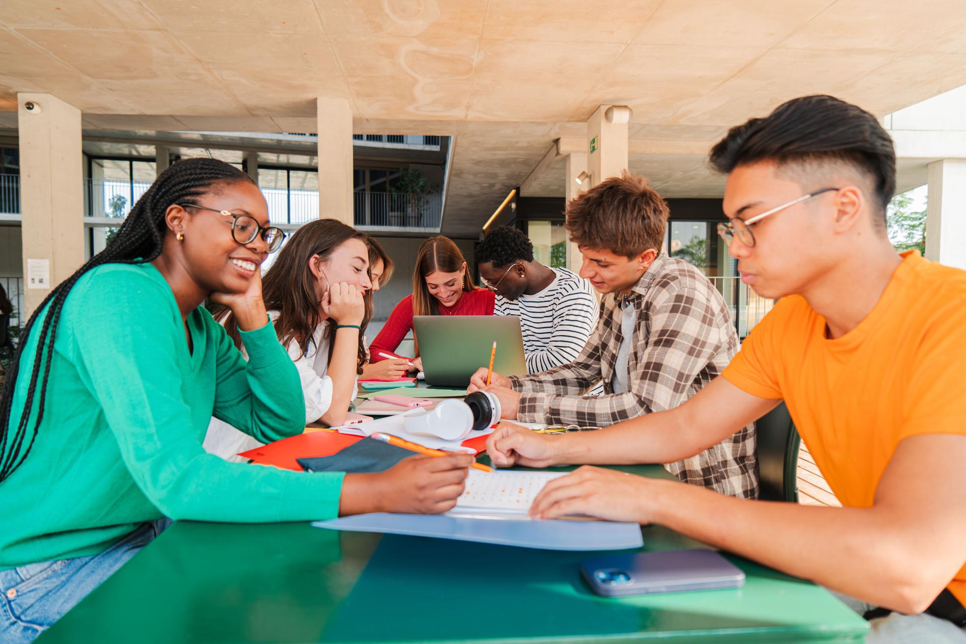 Happy multiracial group of high school students working together, sitting at library with notebooks, writing and using a laptop to search information. Young teenagers studying exams at university